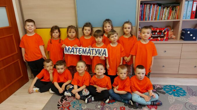 Grupka przedszkolaków w czerwonych strojach. Trzymają duży napis "MATEMATYKA".