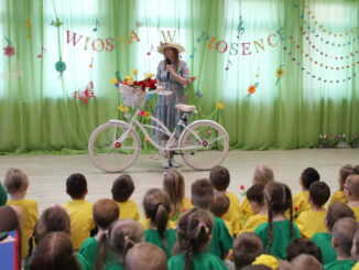 Przed dziećmi stoi Pani Wiosna, z różowym miejskim rowerem. Na kierownicy kosz kwiatów.