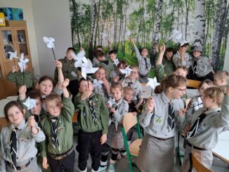 Dzieci w mundurach zuchowych, nad głowami trzymają papierowe wiatraczki. W tle fototapeta z brzozowym zagajnikiem.