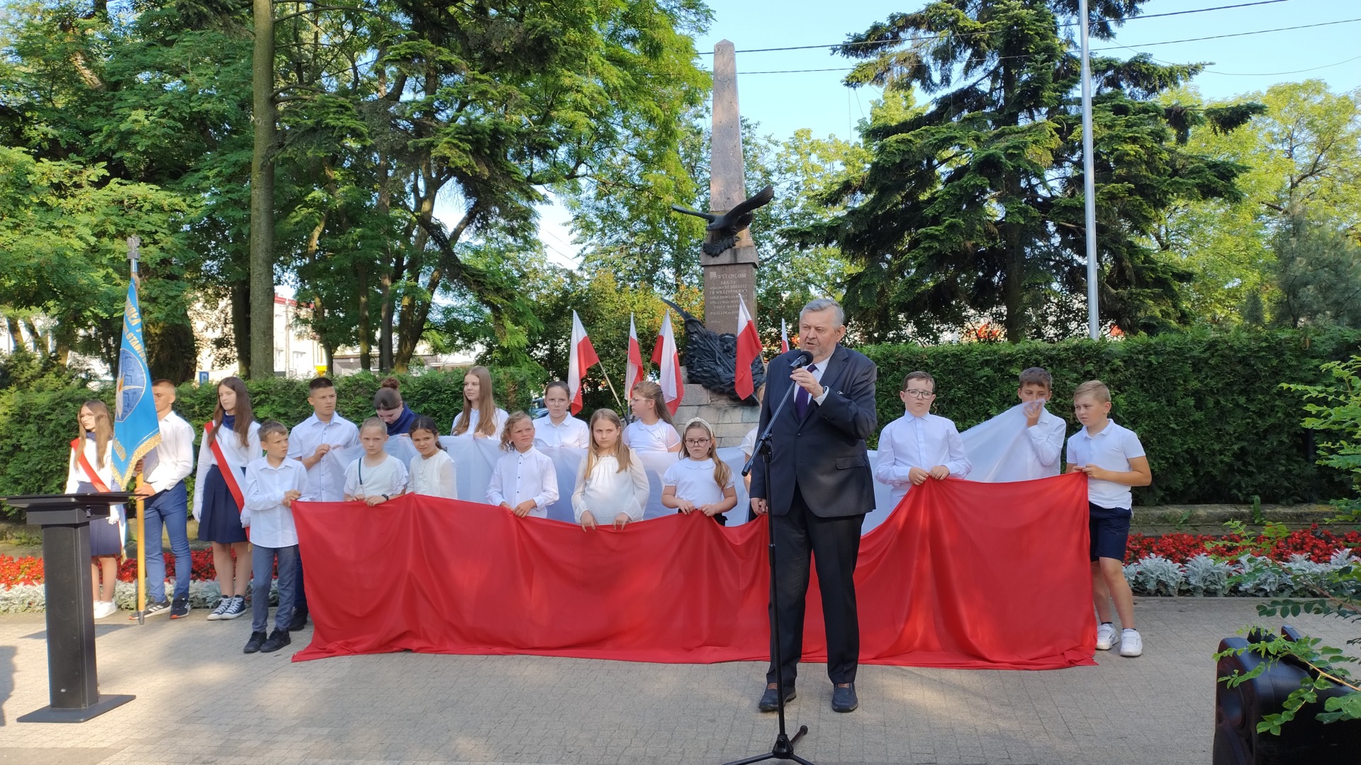 Na pierwszym planie Burmistrz Miasta, dalej dzieci z biało-czerwoną flagą, w tle pomnik ks. Brzóski