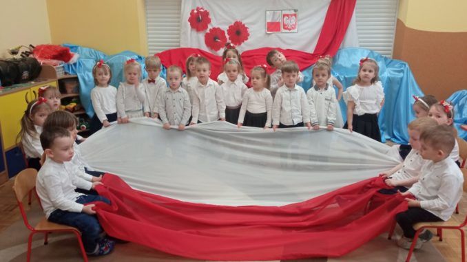 Na górze na środku jest flaga Polski przyozdobiona kwiatami, niżej w półkole stoją przedszkolaki, które trzymają flagę