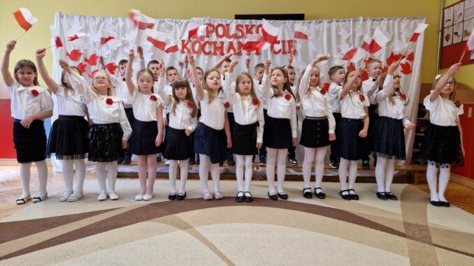Grupa dziewcząt macha biało-czerwonymi chorągiewkami. W tle dekoracja z napisem "Polsko Kocham Cię".