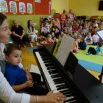 Aneta Mroczek przy elektronicznym pianinie, z przedszkolakiem na kolanach. W tle przedszkolaki siedzące na podłodze.