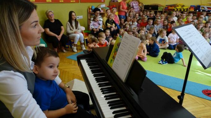 Aneta Mroczek przy elektronicznym pianinie, z przedszkolakiem na kolanach. W tle przedszkolaki siedzące na podłodze.