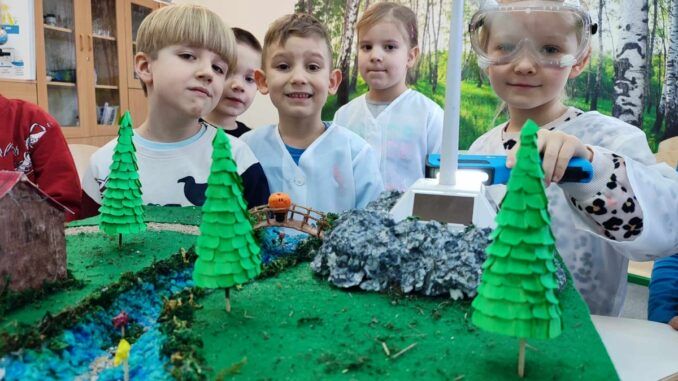 Dzieci na tle fototapety z lasem. Na pierwszym planie makieta terenu zielonego ze zbiornikiem wodnym.