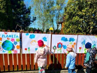 Dzieci malują na dworze, na arkuszach przypiętych do płotu przedszkola.