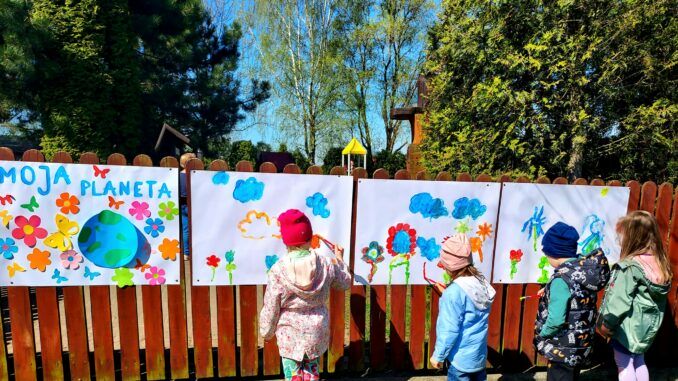 Dzieci malują na dworze, na arkuszach przypiętych do płotu przedszkola.