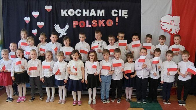 przedszkolaki z biało-czerwonymi chorągiewkami na tle napisu "Kocham Cię Polsko"