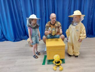 Dwoje dzieci wraz z prowadzącym warsztaty, obok makiety ula. Dzieci ubrane w stroje pszczelarskie.