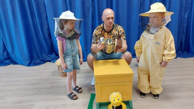 Dwoje dzieci wraz z prowadzącym warsztaty, obok makiety ula. Dzieci ubrane w stroje pszczelarskie.