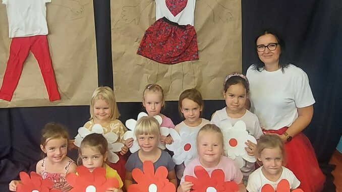 Grupa dzieci wraz z pania, na podłodze. W rekach trzymają papierowe kwiaty. W tle dekoracja wykonana podczas zajęć - chłopiec i dziewczynka w biało-czerwonych ubraniach.