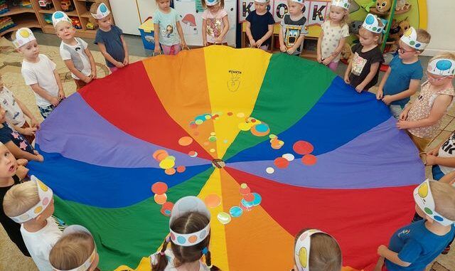 Przedszkolaki trzymają koło z materiału, złożone z klinów w różnych kolorach. Na środku leżą kolorowe kropki.