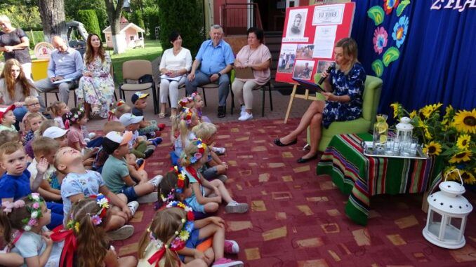 Dzieci podczas czytania, pod budynkiem przedszkola. W tle dorośli uczestnicy, z Burmistrzem Miasta.