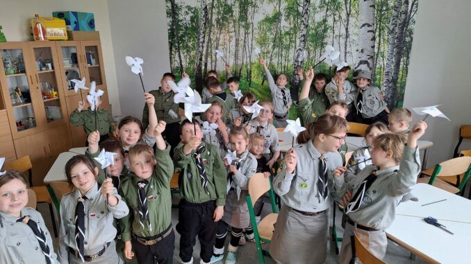 Dzieci w mundurach zuchowych, nad głowami trzymają papierowe wiatraczki. W tle fototapeta z brzozowym zagajnikiem.