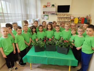 Dzieci w zielonych koszulkach stoją w półokręgu. Na środku stół, a na nim skrzynki z kwiatami.