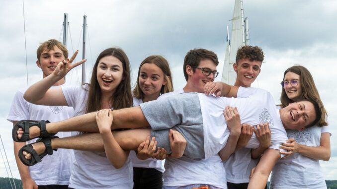 Grafika promocyjna programu: grupa młodzieży trzyma na rękach kolegę w pozycji horyzontalnej. W tle widok na jezioro.